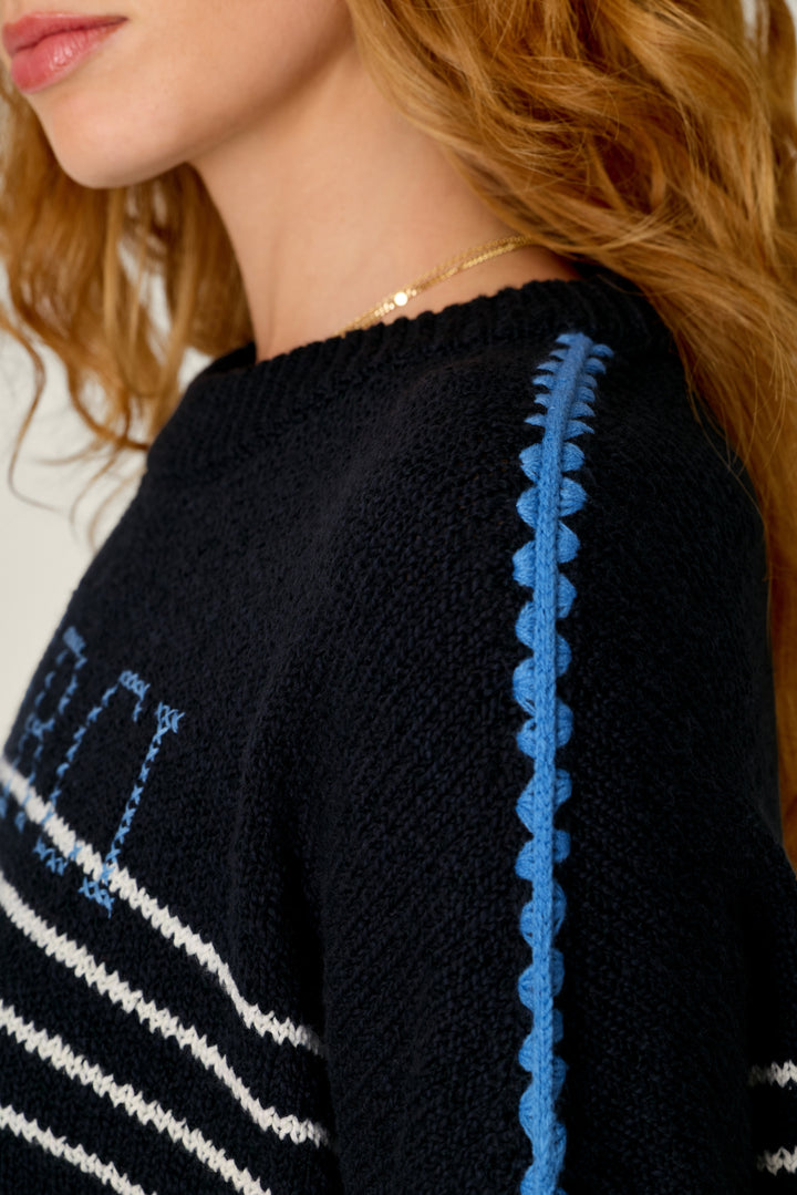 Close-up of a person wearing a black knit sweater with blue and white striped details and a blue scalloped trim running down the shoulder.