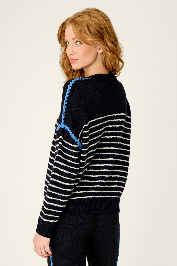 A woman with red hair is shown from a side-back angle wearing a black knit sweater with white horizontal stripes and bright blue accent lines, paired with black pants, against a plain light background.