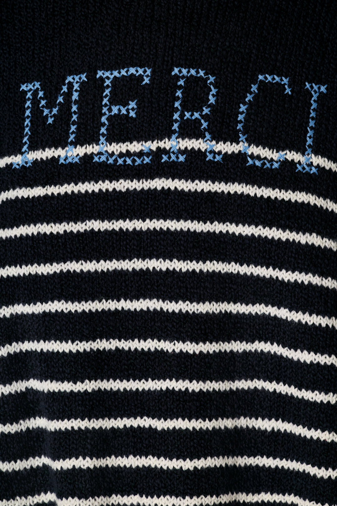 Close-up of a knit sweater featuring horizontal navy and white stripes with the word 'MERCI' knitted in light blue across the chest.