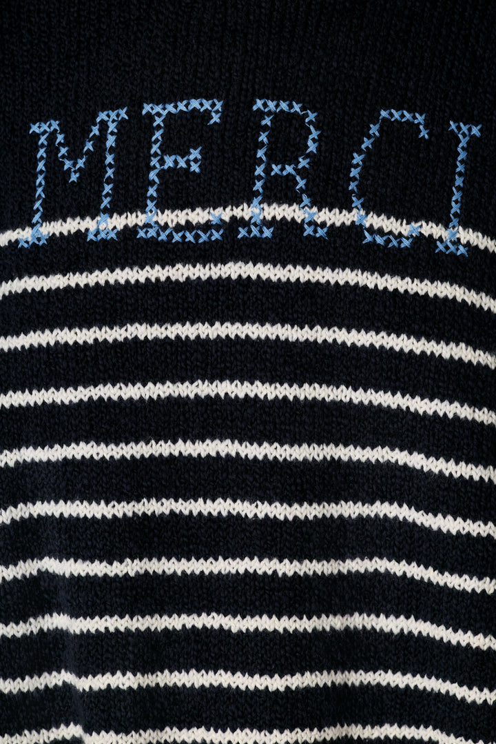 Close-up of a knit sweater featuring horizontal navy and white stripes with the word 'MERCI' knitted in light blue across the chest.