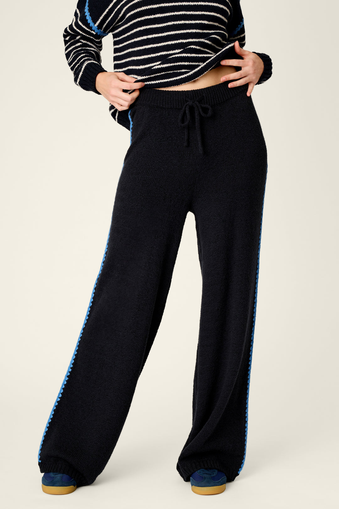 A person wearing black wide-leg knit pants with a blue textured stripe down the sides, paired with a black-and-white striped cropped sweater, against a plain light background.