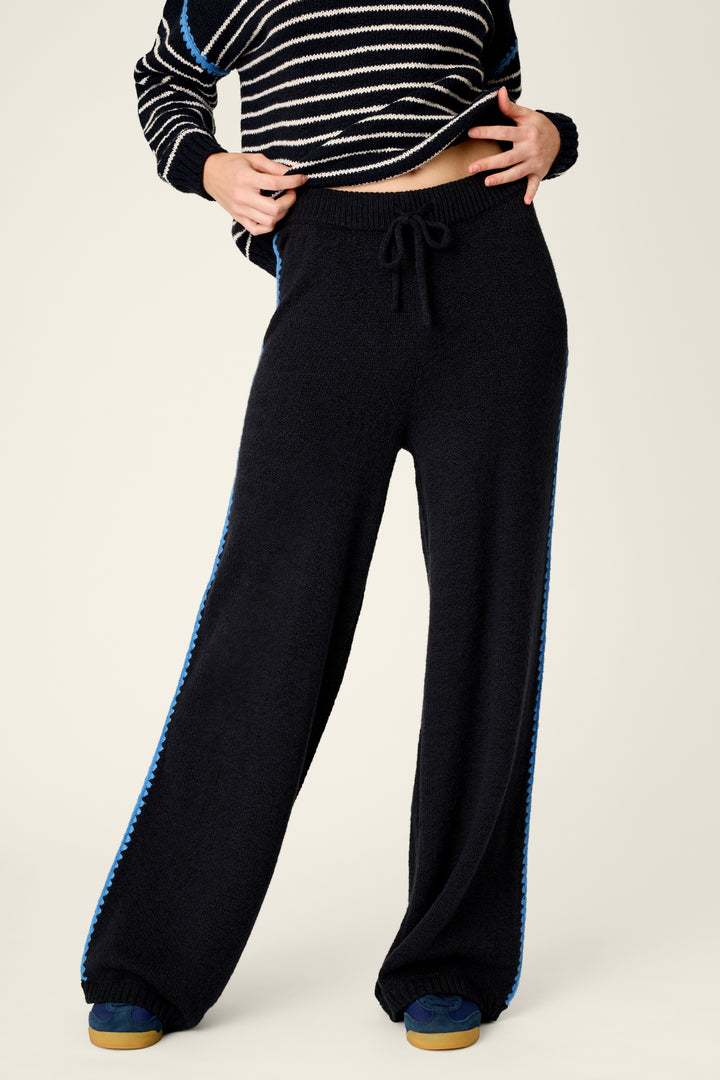 A person wearing black wide-leg knit pants with a blue textured stripe down the sides, paired with a black-and-white striped cropped sweater, against a plain light background.