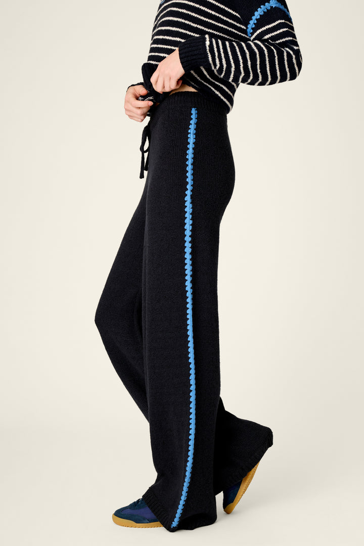 A person wearing black knit wide-leg pants with a bright blue braided stripe down the side, paired with a black-and-white striped sweater and blue shoes, against a plain light beige studio background.