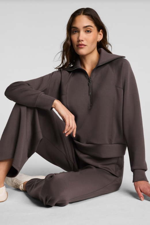 Woman wearing a brown QUARTER ZIP TOP and pants sitting on a light gray background