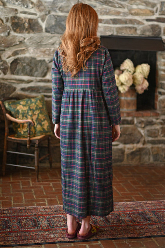 Woman in a plaid nightdress standing in a room with stone walls and a fireplace.