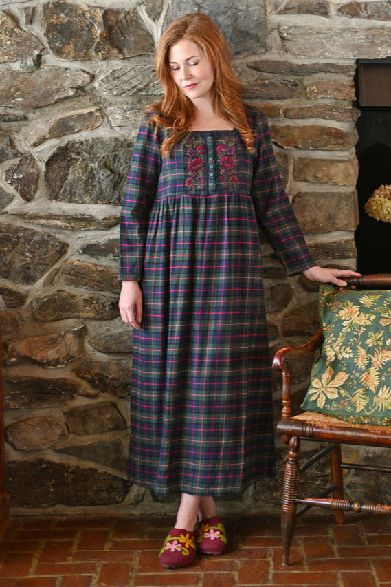 Woman wearing a plaid nightdress with embroidery standing in front of a stone wall.