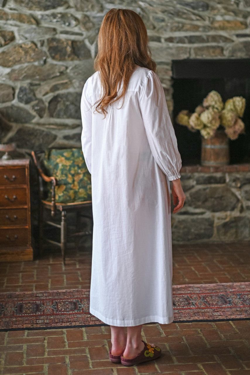 Woman in a white nightdress standing in a room with stone walls and a fireplace.