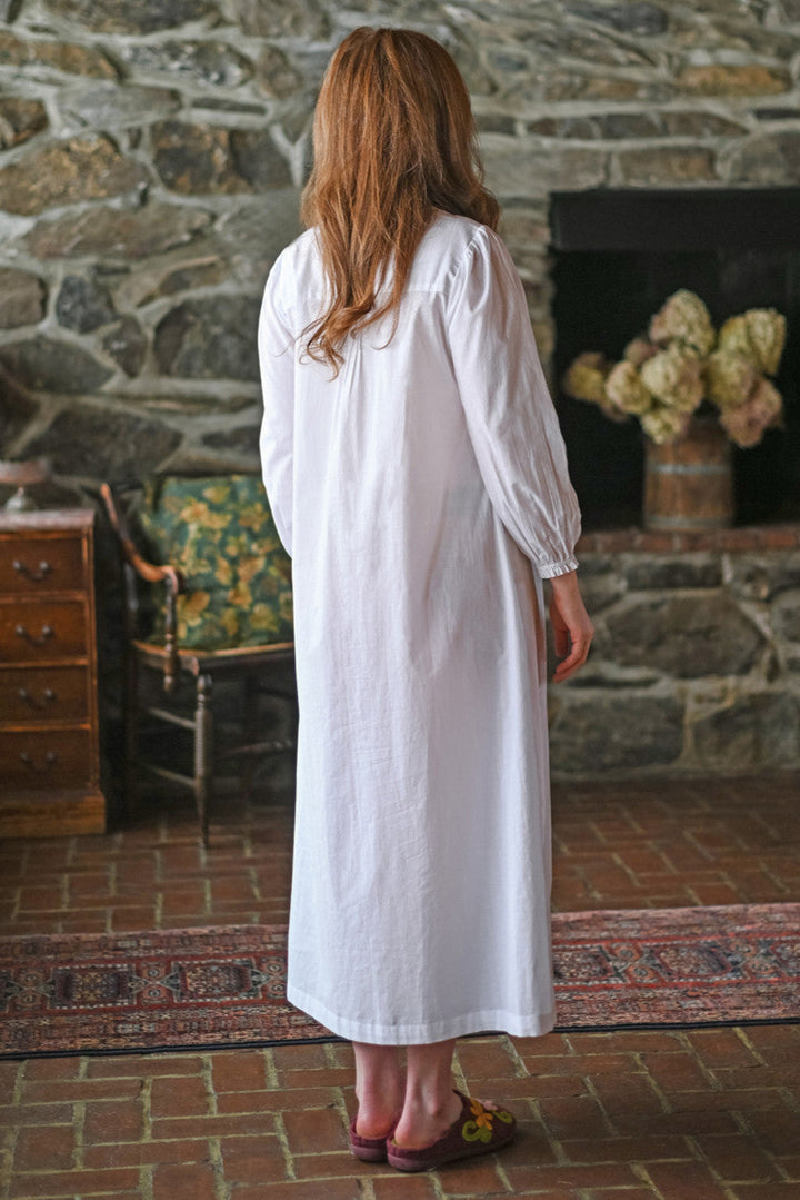 Woman in a white nightdress standing in a room with stone walls and a fireplace.