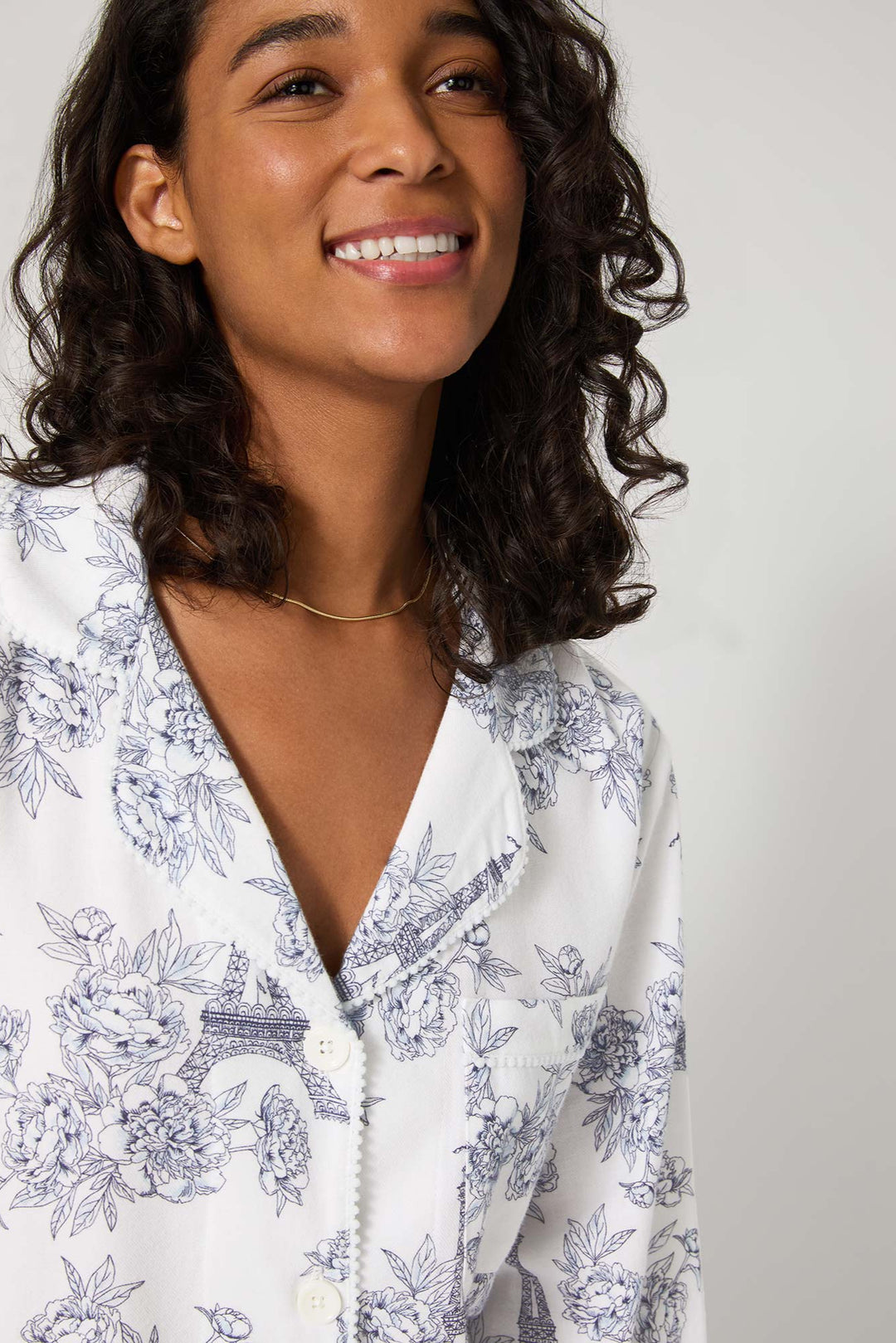 Woman wearing a white pajama with blue floral and architectural print against a plain background