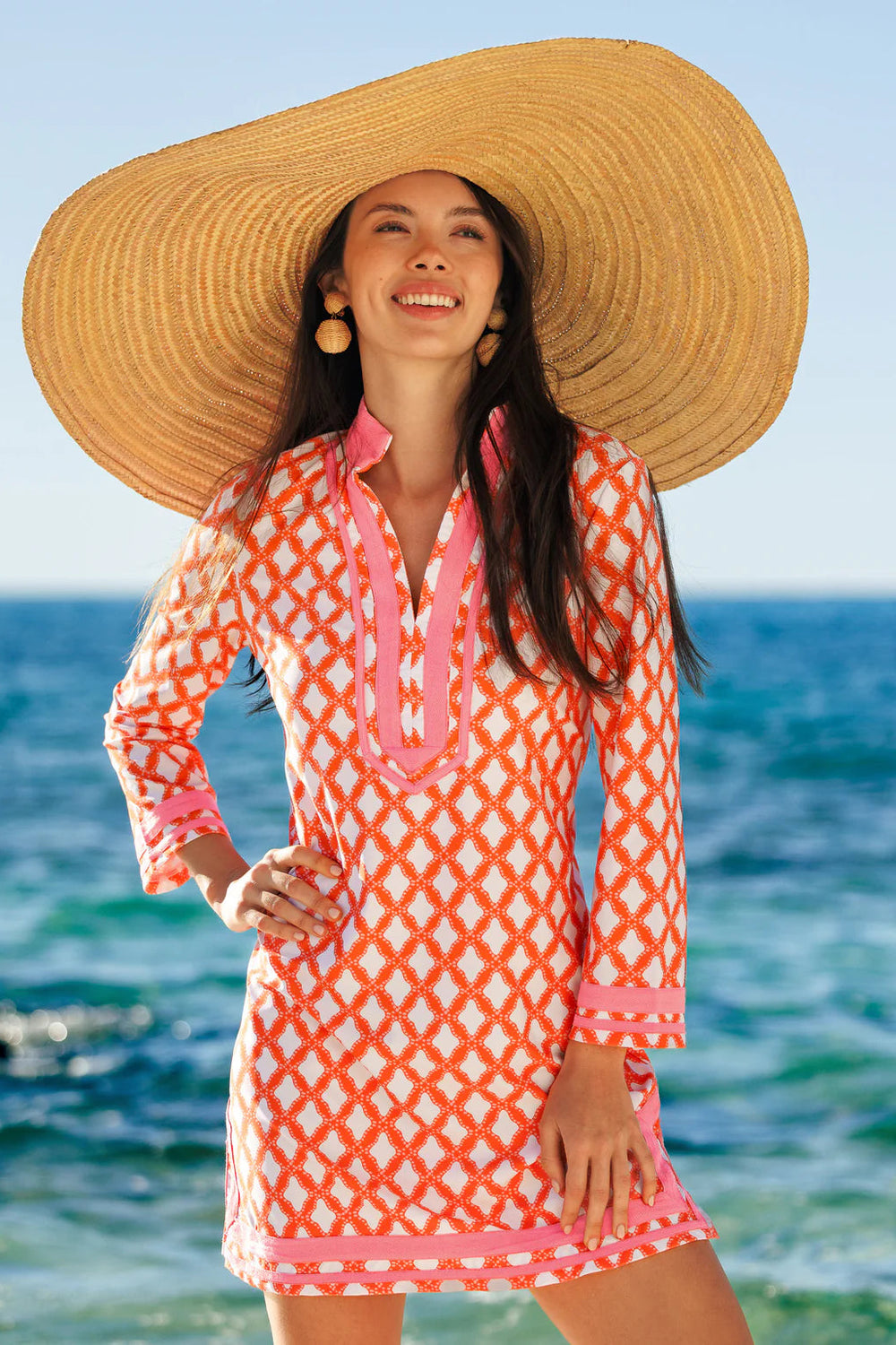Woman in a pink and orange patterned dress with a large straw hat standing by the ocean.