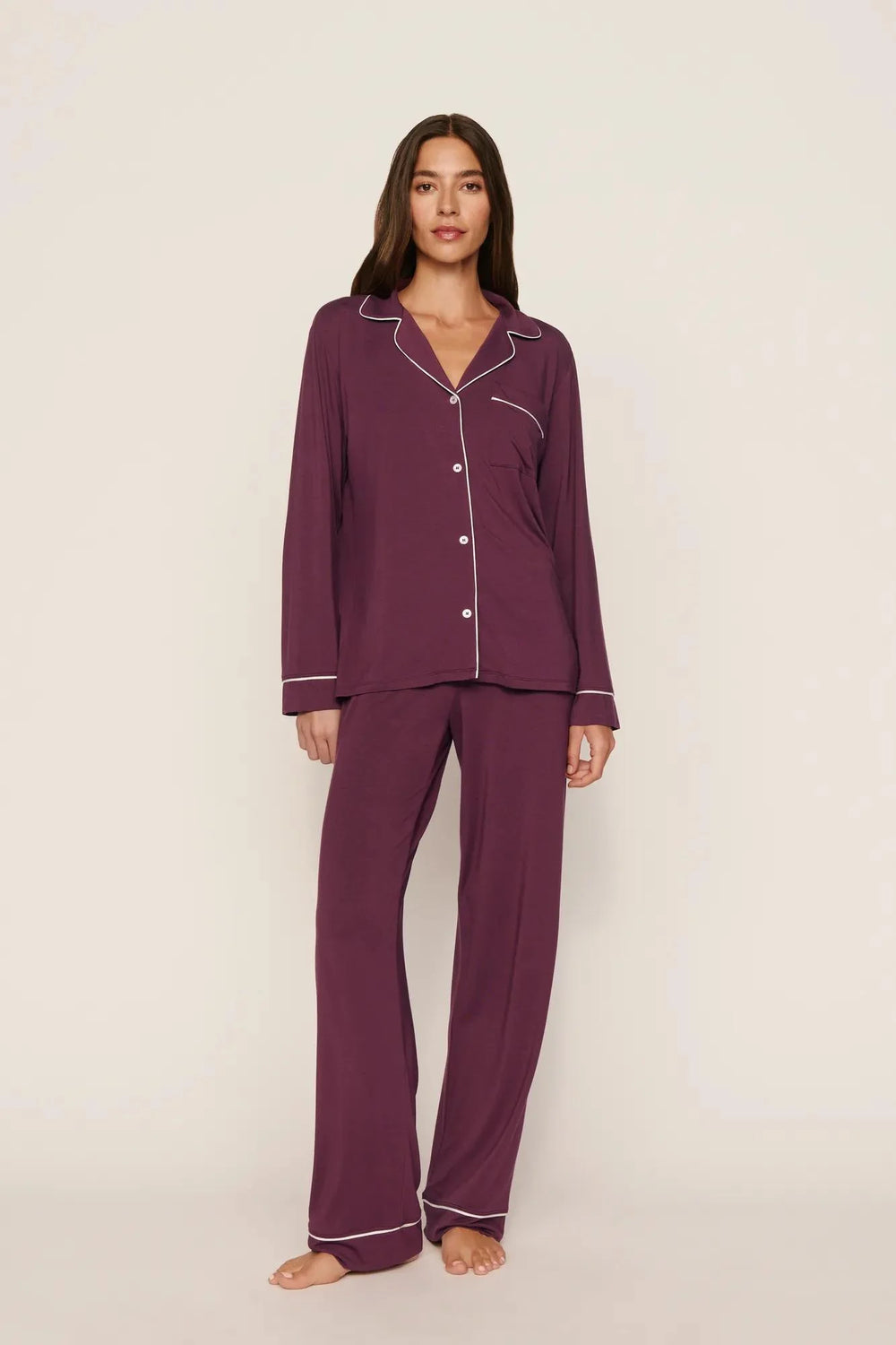 A woman modeling a two-piece pajama set in deep plum purple with white piping trim, standing against a plain light beige studio background.
