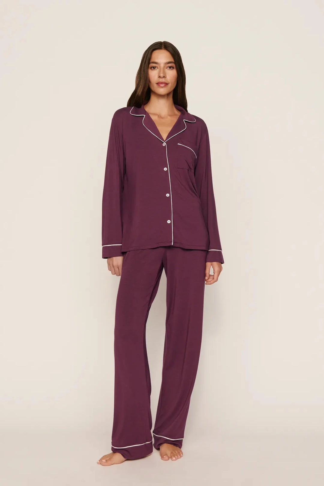 A woman modeling a two-piece pajama set in deep plum purple with white piping trim, standing against a plain light beige studio background.