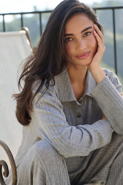 A woman with long dark hair sits on a white outdoor lounge chair, wearing a light gray ribbed long-sleeve button-up shirt and matching gray pants. The background shows a blurred outdoor scene with a railing and greenery.