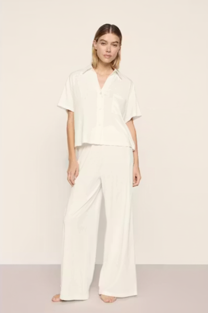 A woman modeling a matching white two-piece lounge set consisting of a short-sleeve button-up shirt and wide-leg trousers, set against a plain off-white studio background.