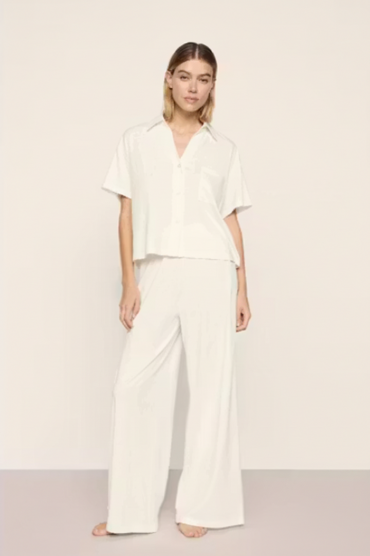 A woman modeling a matching white two-piece lounge set consisting of a short-sleeve button-up shirt and wide-leg trousers, set against a plain off-white studio background.
