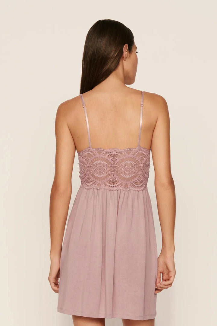 A woman shown from the back wearing a dusty pink nightgown with thin straps and a lace bodice, photographed against a plain light beige studio background.