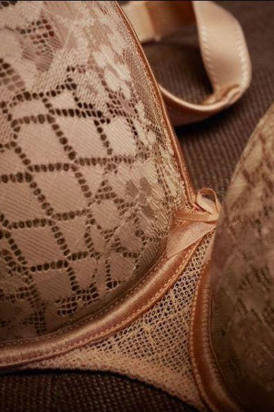 Close-up of a beige lace bra with a satin trim and diamond-patterned lace cups, resting on a dark brown textured surface.
