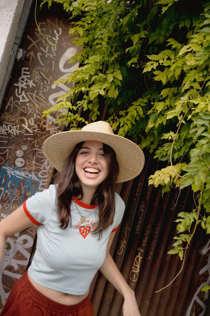 A woman wearing a straw wide-brim hat, a light blue ringer t-shirt with red trim and a heart graphic, and a rust-colored skirt, smiling outdoors against a background of green ivy and a graffiti-covered wall.