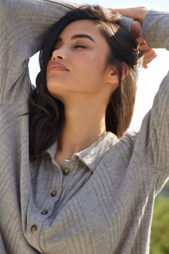 A woman wearing a light gray ribbed long-sleeve button-up shirt, stretching her arms above her head in an outdoor setting with bright sunlight.