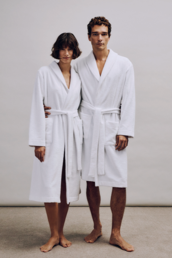 Two people wearing white terry robes standing against a plain background