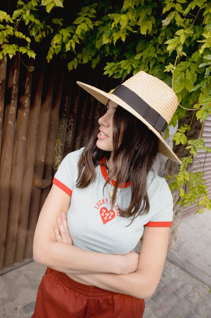 A woman wearing a straw sun hat with a black band, a light blue ringer t-shirt with red trim and a red heart graphic, and rust-colored shorts, standing outdoors under green vines.