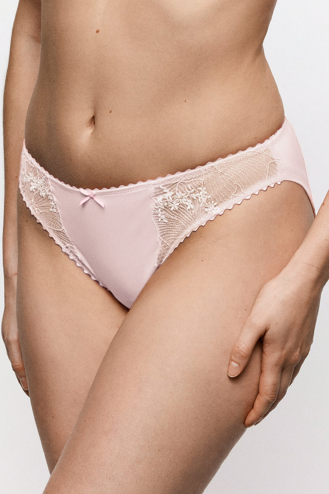 A close-up of a model wearing light pink lace-trimmed underwear against a plain light gray studio background.