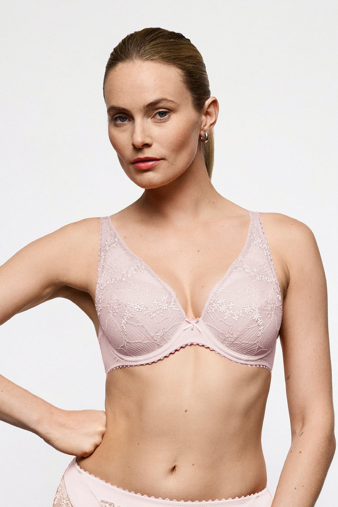 A woman modeling a light pink lace bra and matching high-waisted briefs against a plain light gray studio background.