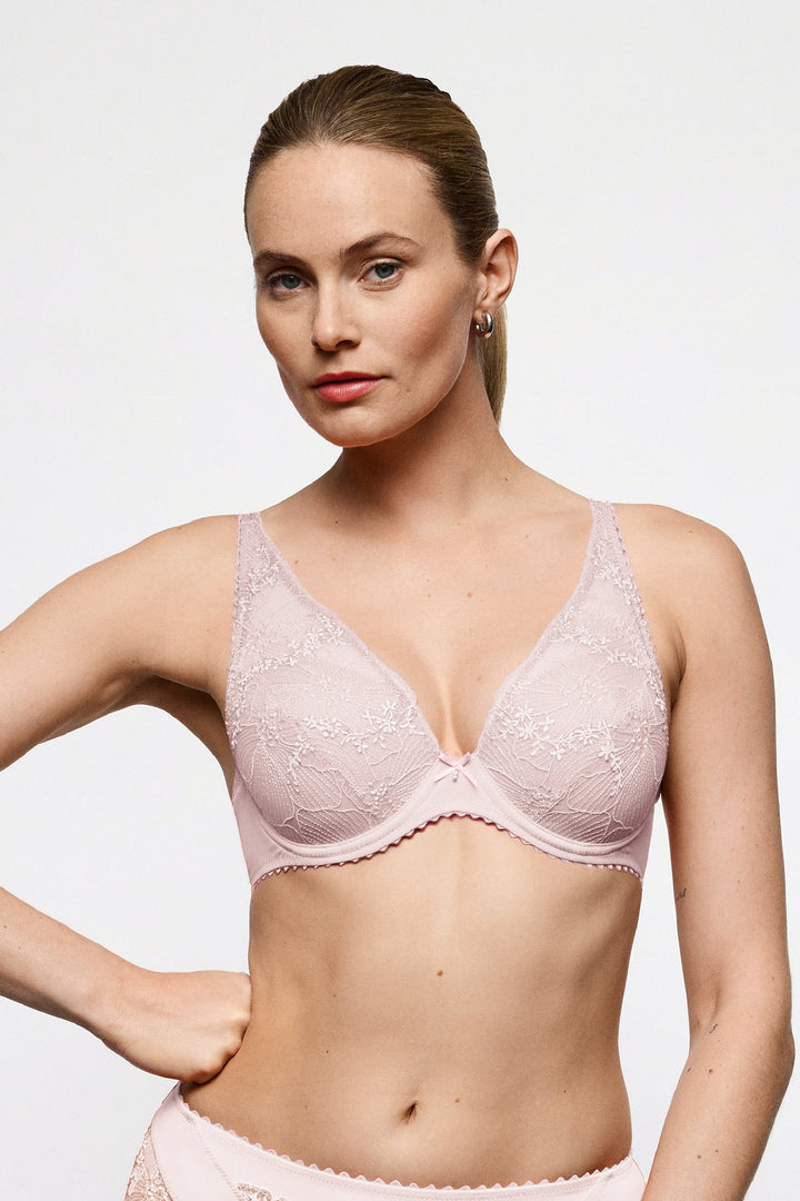 A woman modeling a light pink lace bra and matching high-waisted briefs against a plain light gray studio background.
