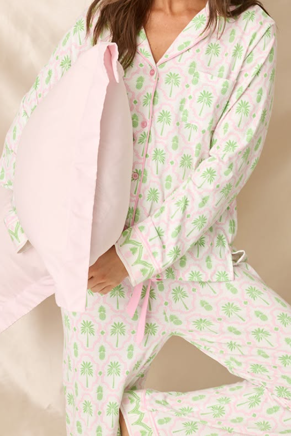 A person wearing a light pink pajama set with a green tropical print, holding a matching light pink pillow against a soft beige fabric backdrop.