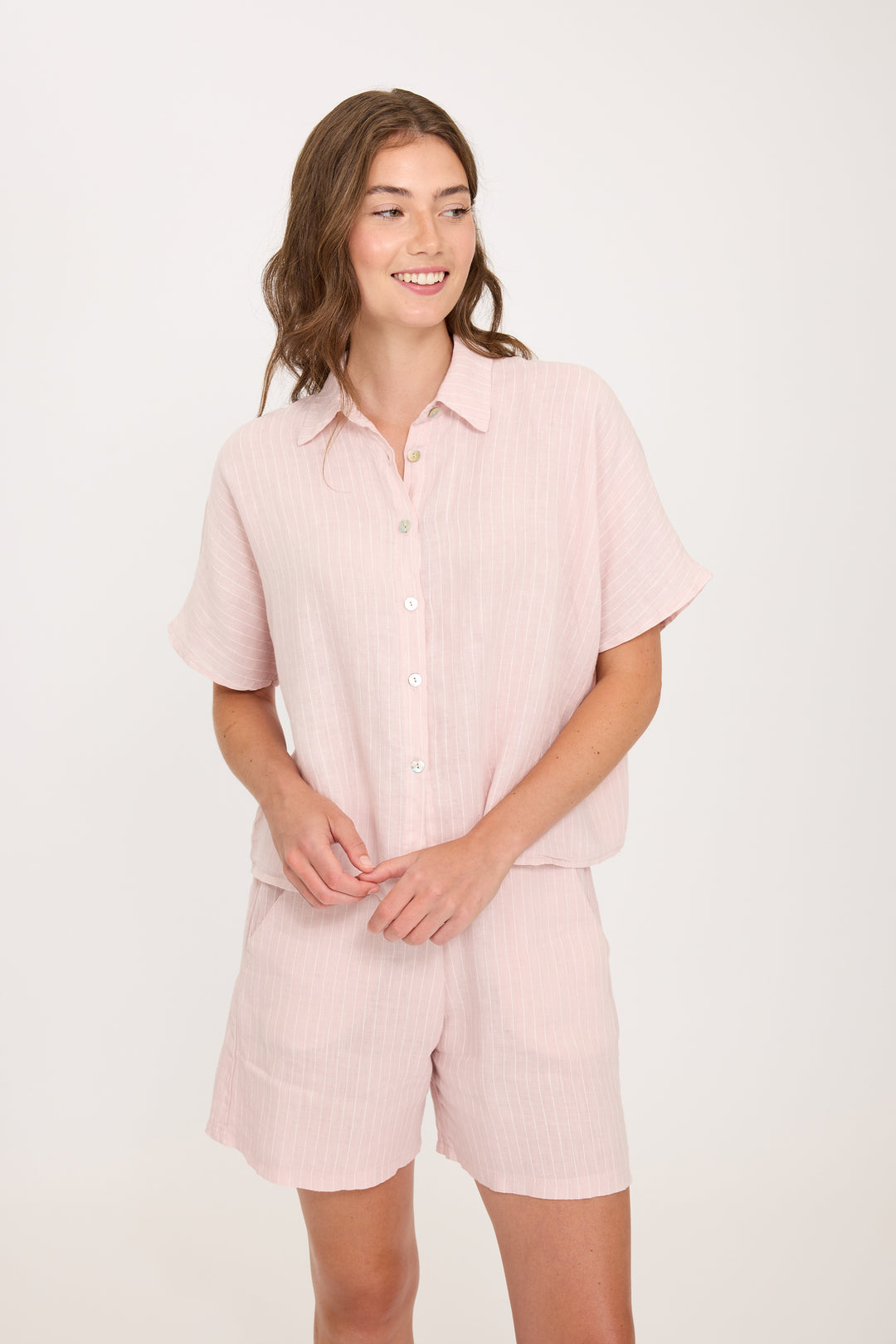 Woman wearing a light pink striped linen shirt and shorts set on a white background. Vacation outfit, perfect for summertime or hot weather.