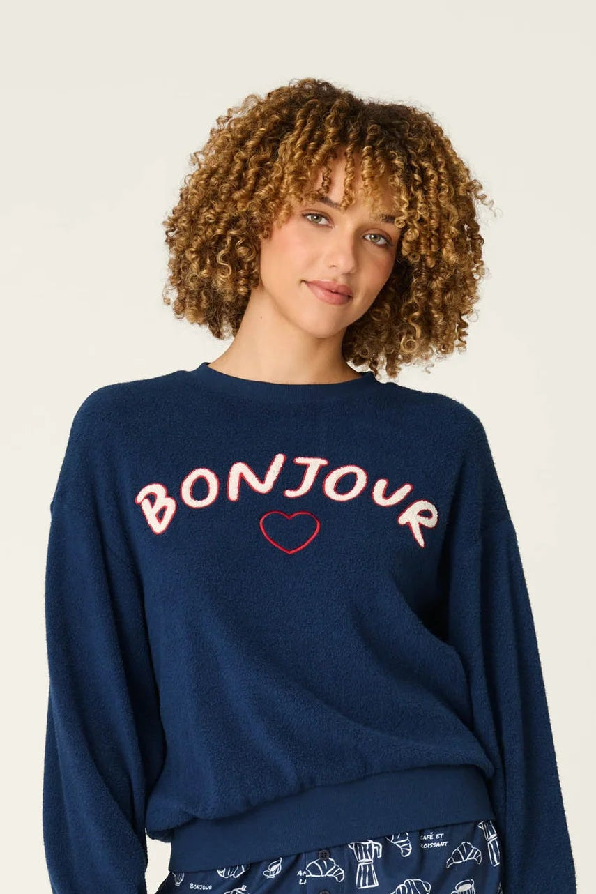 A woman modeling a navy blue pajama set consisting of a sweatshirt with 'BONJOUR' printed in white and a small pink heart, and matching patterned shorts, against a plain light background.