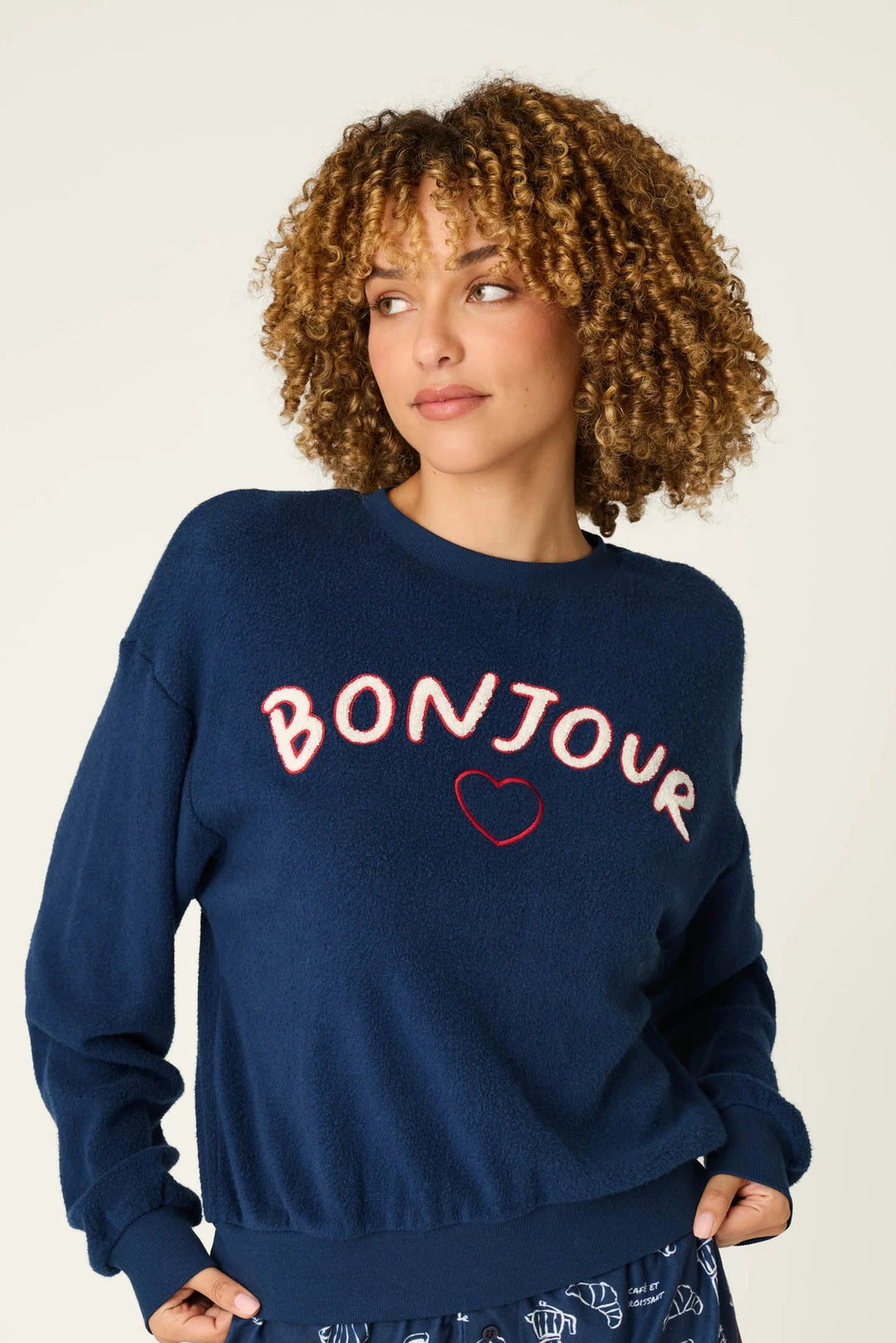 A woman modeling a navy blue sweatshirt and matching shorts set. The sweatshirt features 'BONJOUR' in red letters with a small red heart outline.