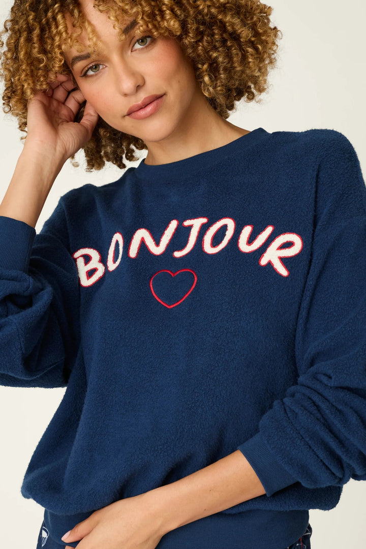 A woman wearing a navy blue sweatshirt with 'BONJOUR' and a heart graphic in white and red, paired with matching patterned shorts, against a plain light background.
