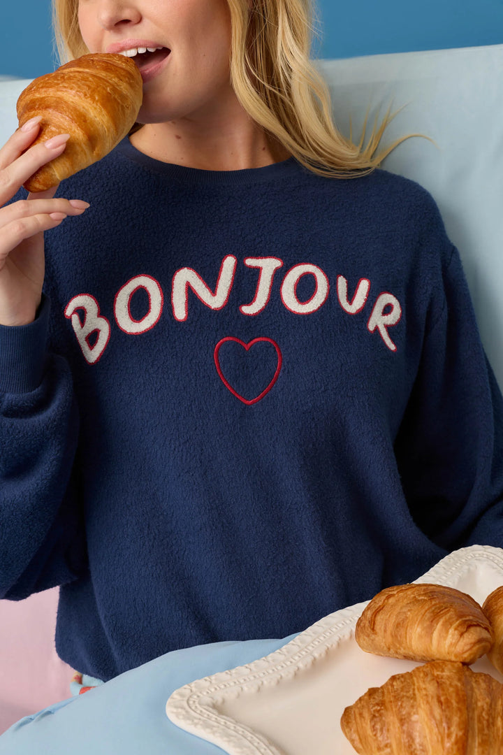 A woman wearing a navy blue fuzzy sweater with 'BONJOUR' and a red heart embroidered on the front, eating a croissant in a cozy indoor setting.