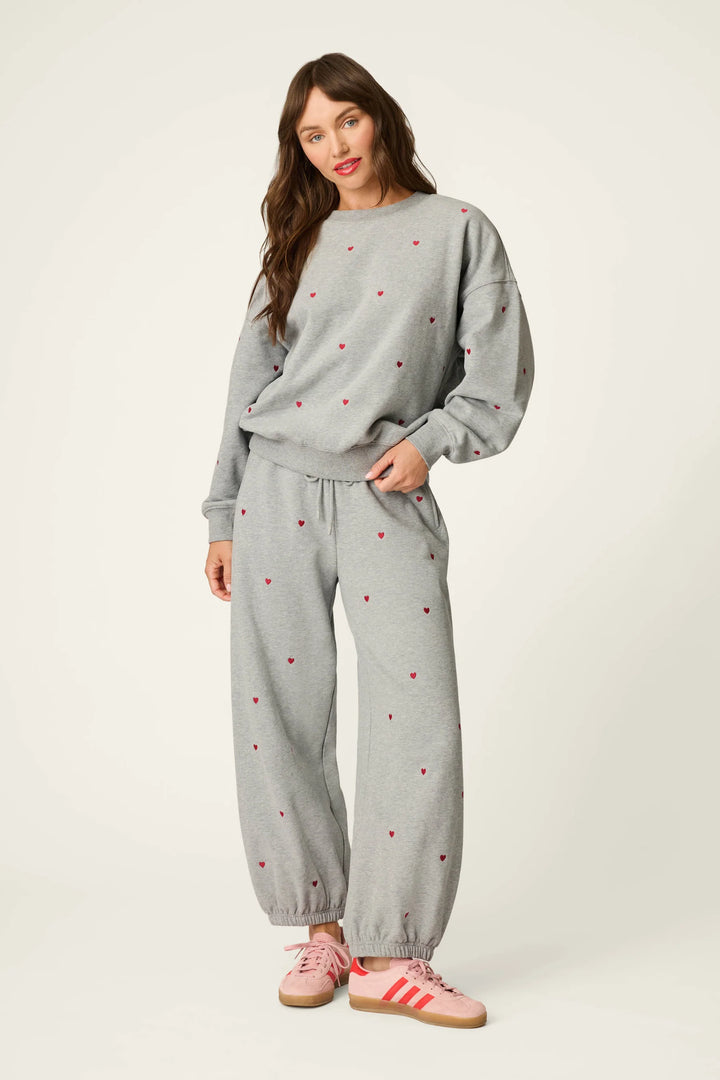 A woman modeling a light gray sweatshirt and matching jogger pants set with small red embroidered dots, styled with pink sneakers against a plain off-white studio background.