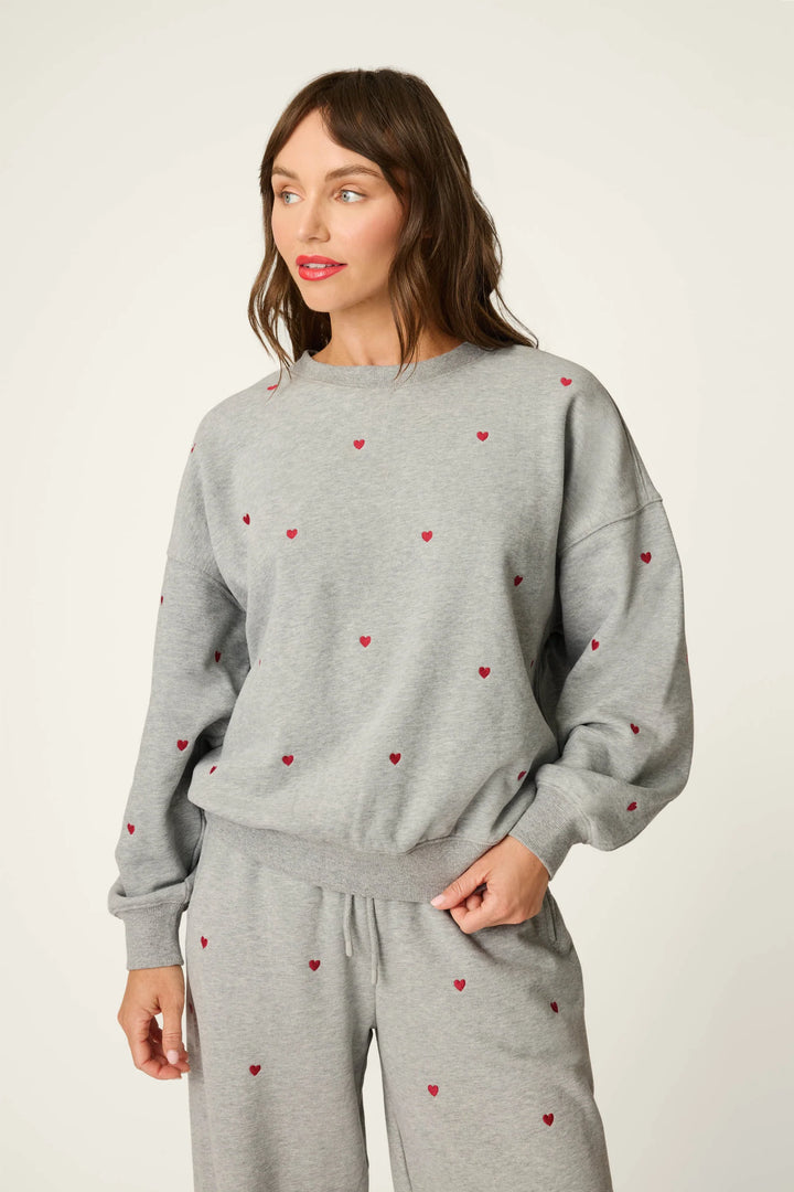 A woman modeling a light gray sweatshirt and matching sweatpants set, both decorated with small red embroidered hearts, against a plain off-white studio background.