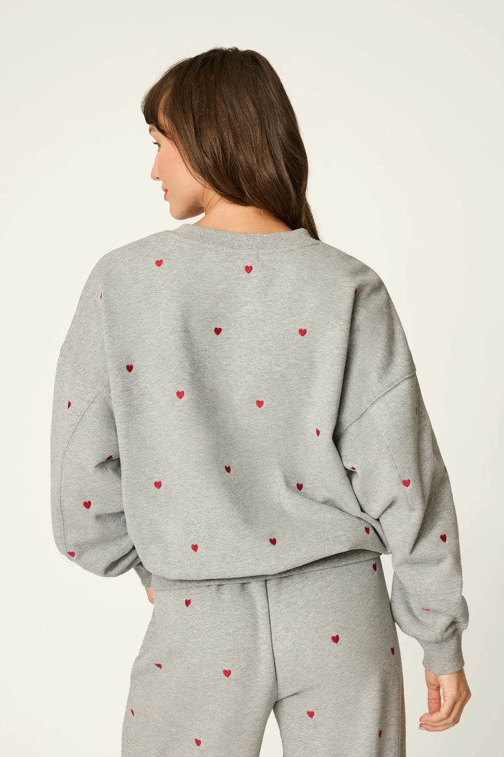A woman shown from the back wearing a light gray heather sweatshirt and matching sweatpants, both decorated with a small red heart pattern, against a plain off-white studio background.