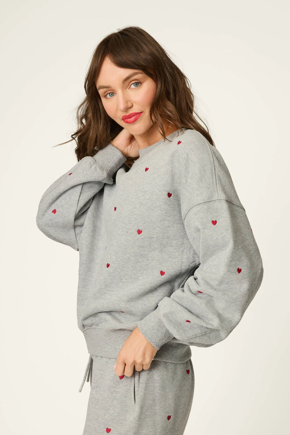 A woman modeling a light gray heather sweatshirt and matching sweatpants set, decorated with small red embroidered hearts, against a plain off-white studio background.