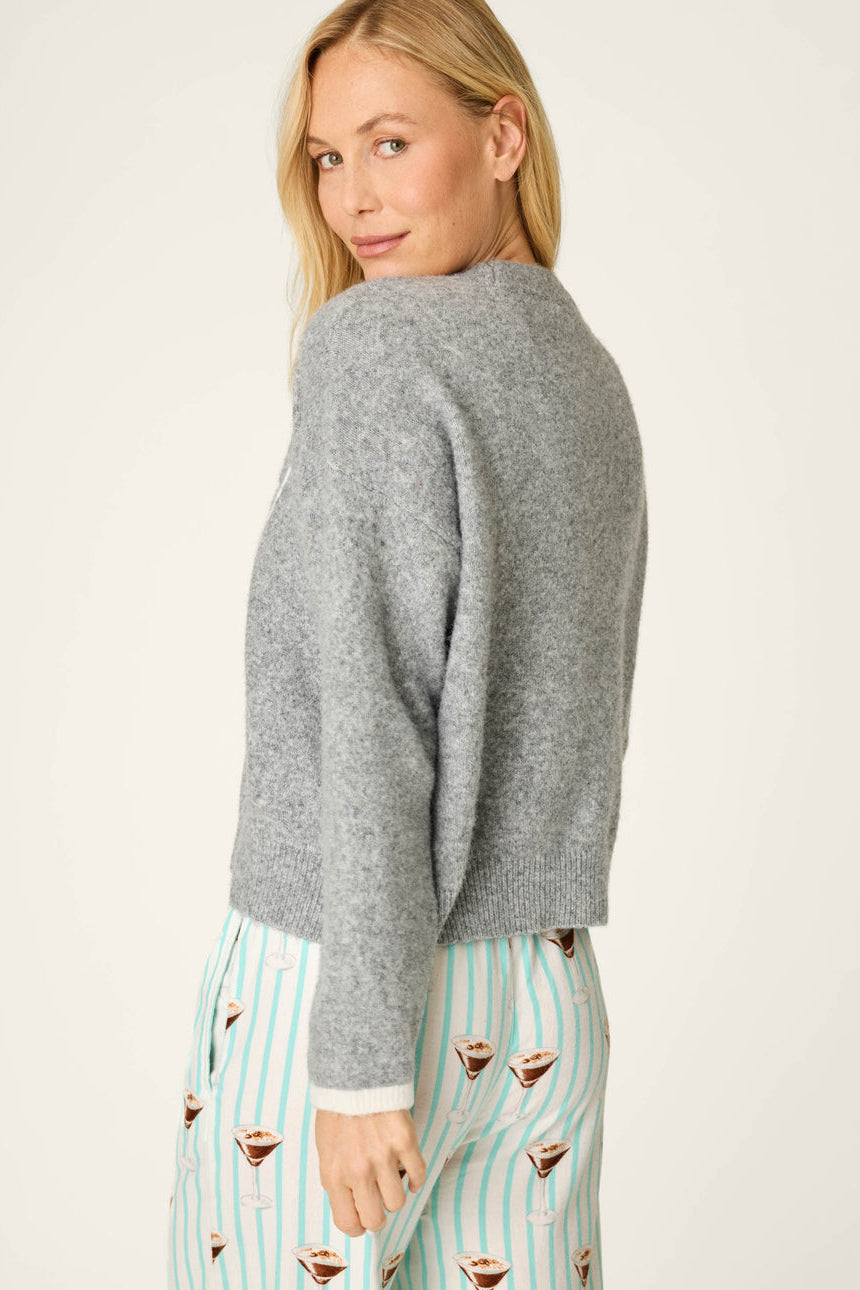 A woman shown from the back wearing a light gray knit sweater and mint green and white striped pajama pants with cocktail prints, against a plain off-white studio background.