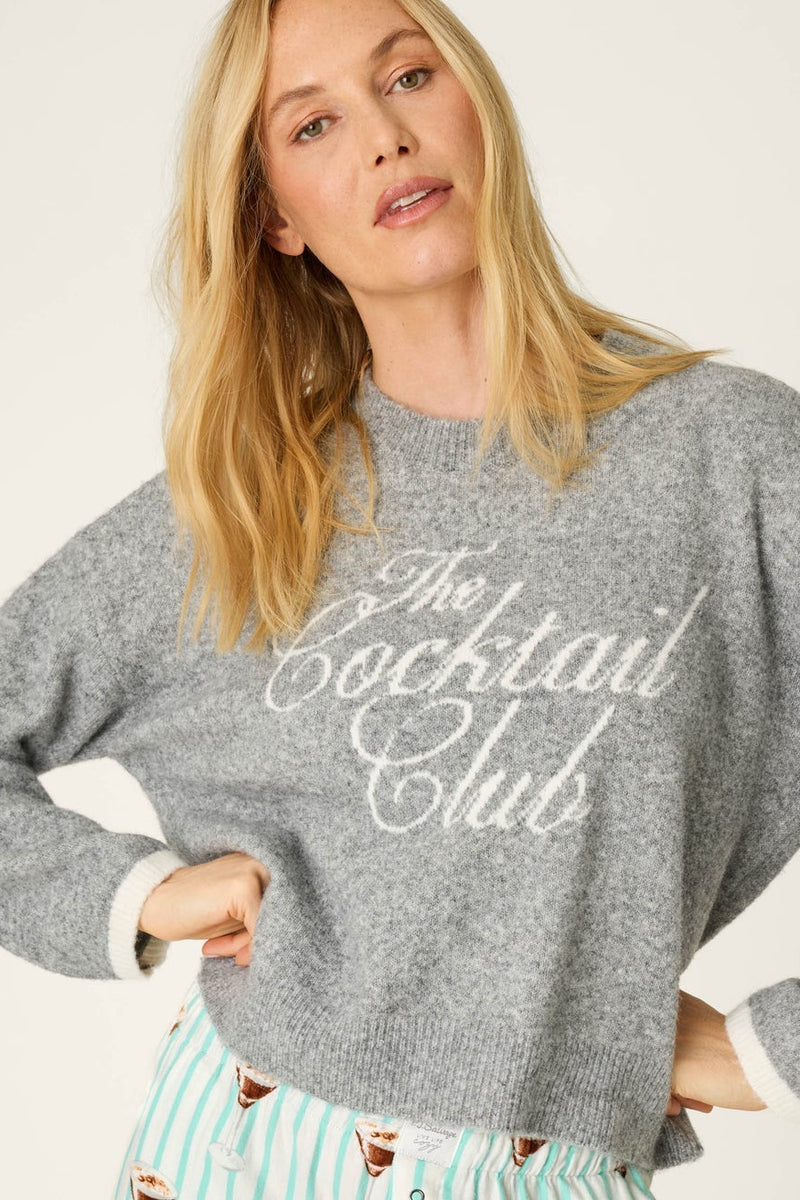 A woman wearing a gray knit sweater with 'The Cocktail Club' written across the chest in white script, paired with mint green and white striped pajama pants featuring a cocktail print.
