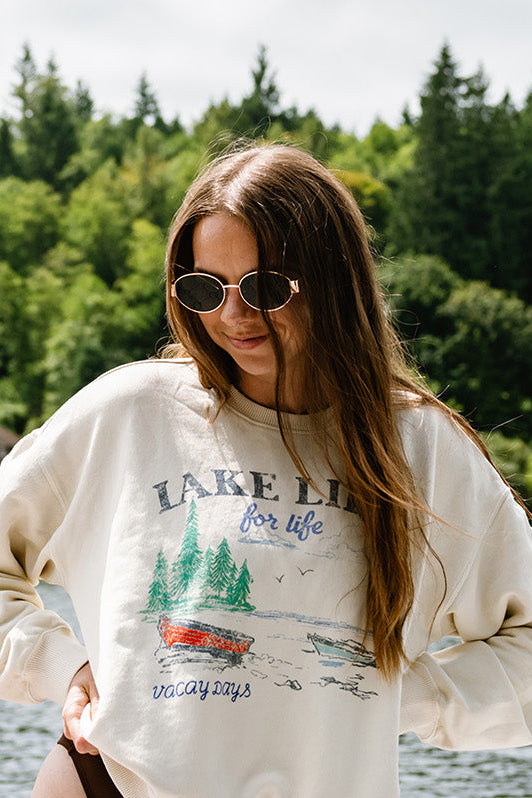 Woman wearing a 'Lake Life' sweatshirt by a lake with trees and houses in the background