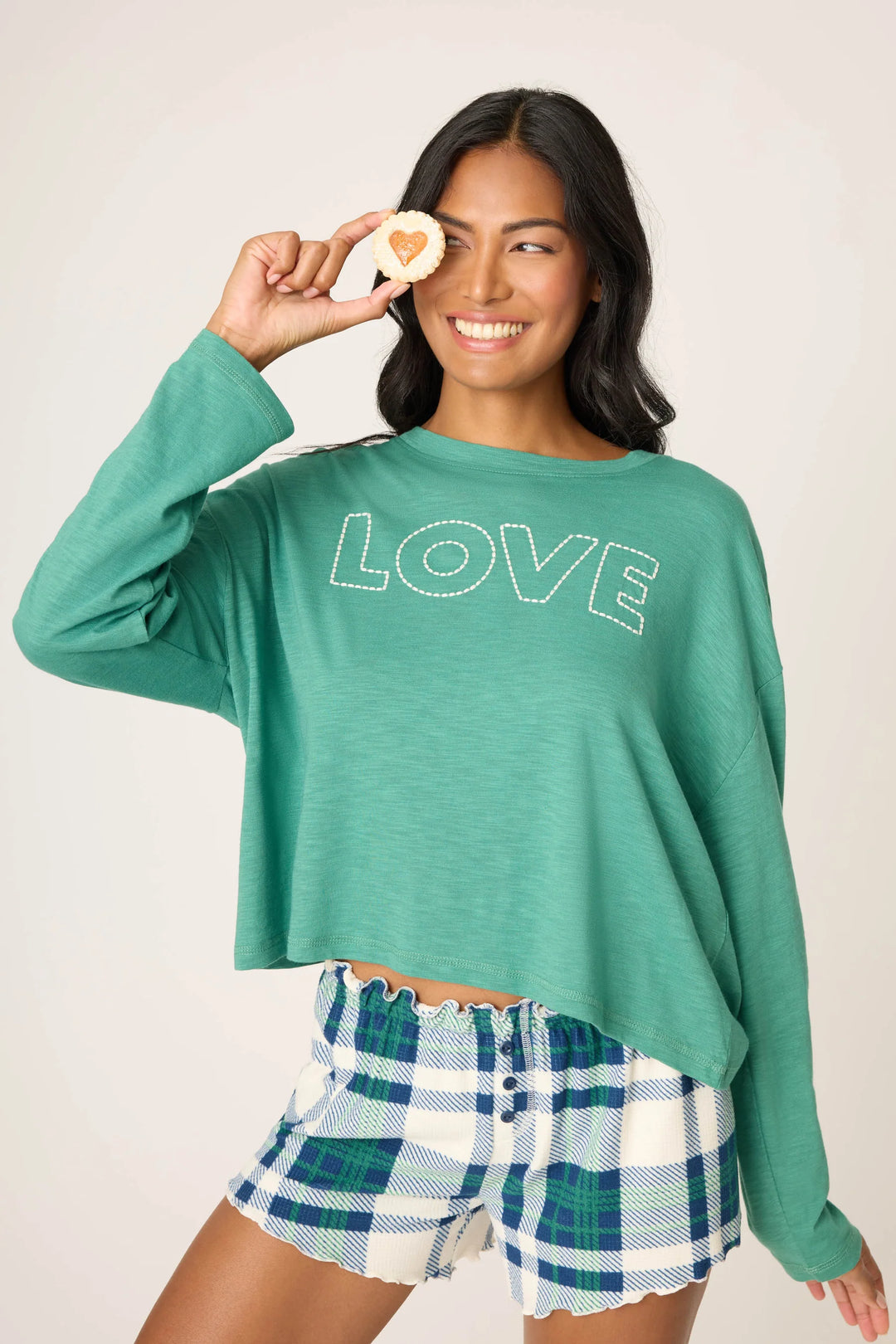 Woman wearing a green 'LOVE' sweatshirt and plaid shorts, holding a cookie with a heart on it.