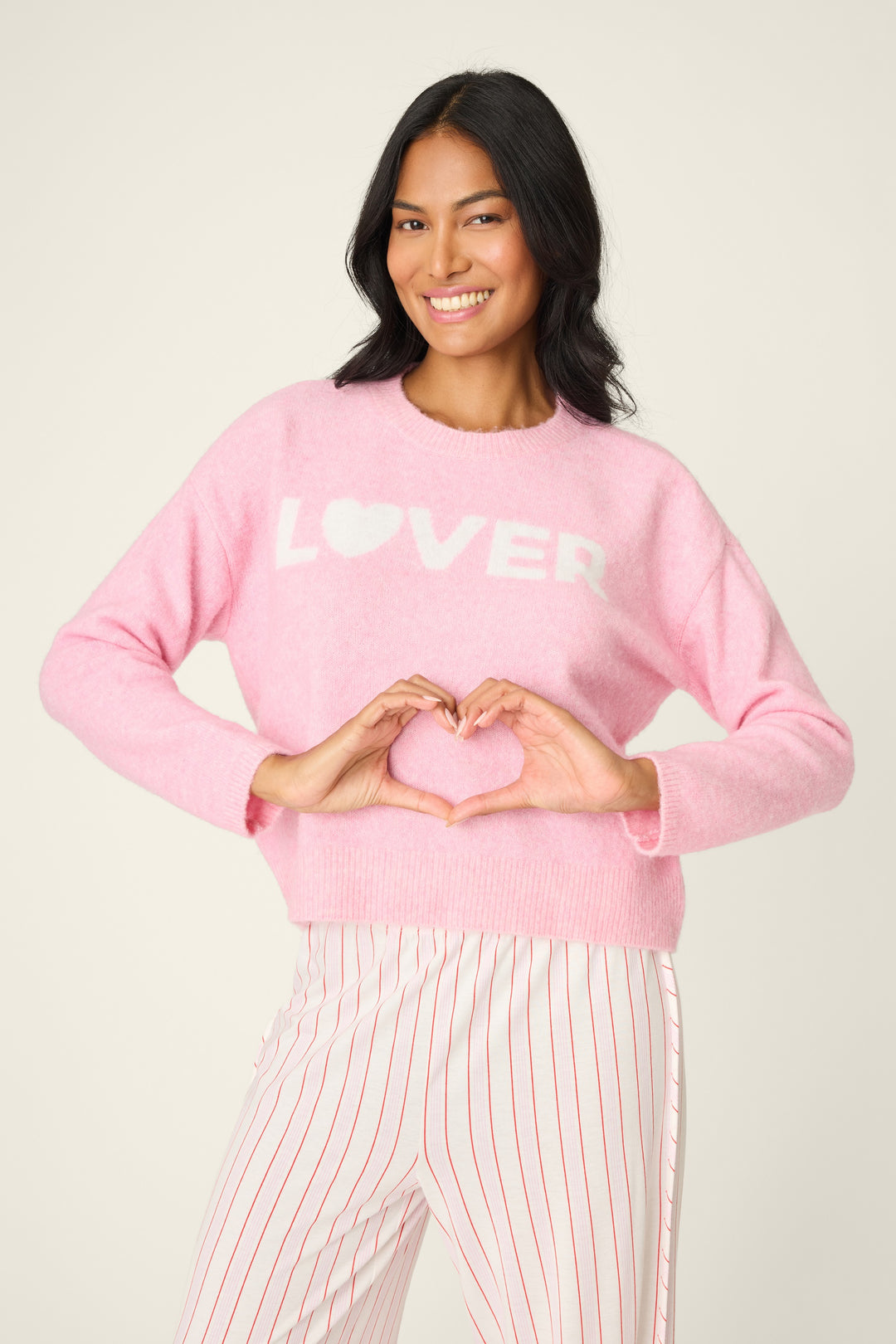 Woman wearing a pink sweater with 'LOVER' text, making a heart shape with her hands on a light background