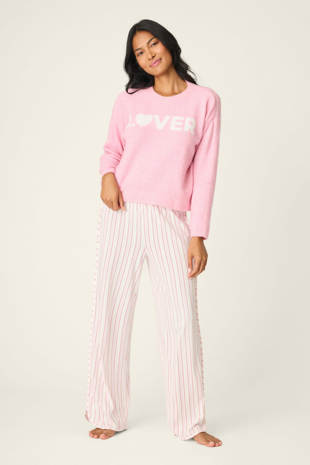 Woman wearing a pink sweatshirt with 'LOVER' printed on it and striped pants against a white background