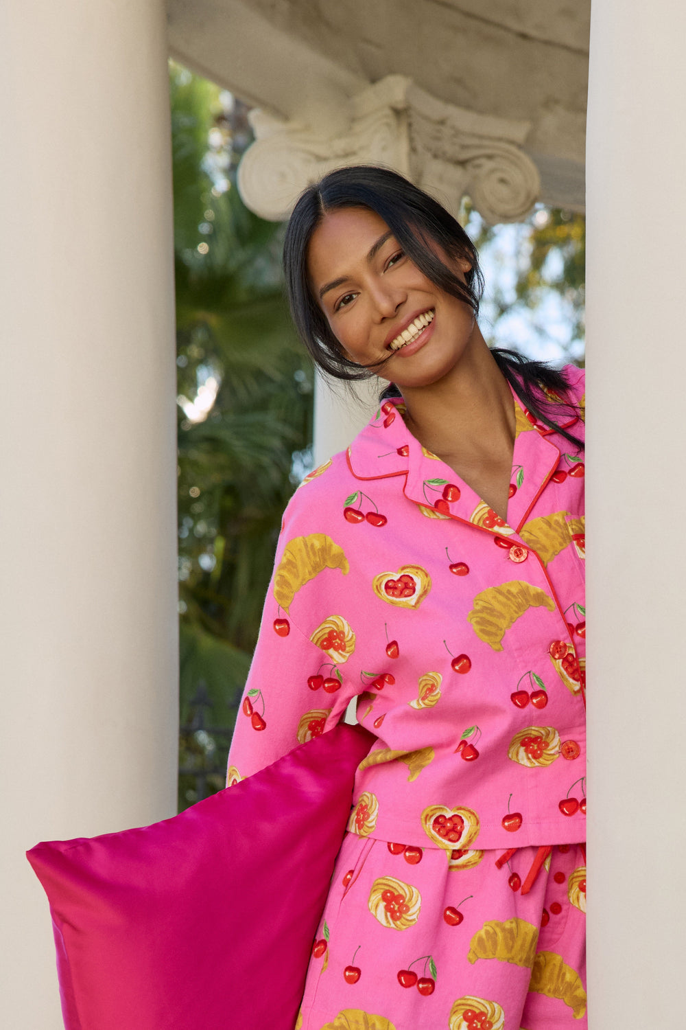 A woman wearing a bright pink pajama set with a cherry and fruit print, holding a matching hot pink pillow in a sunlit outdoor corridor framed by white columns.