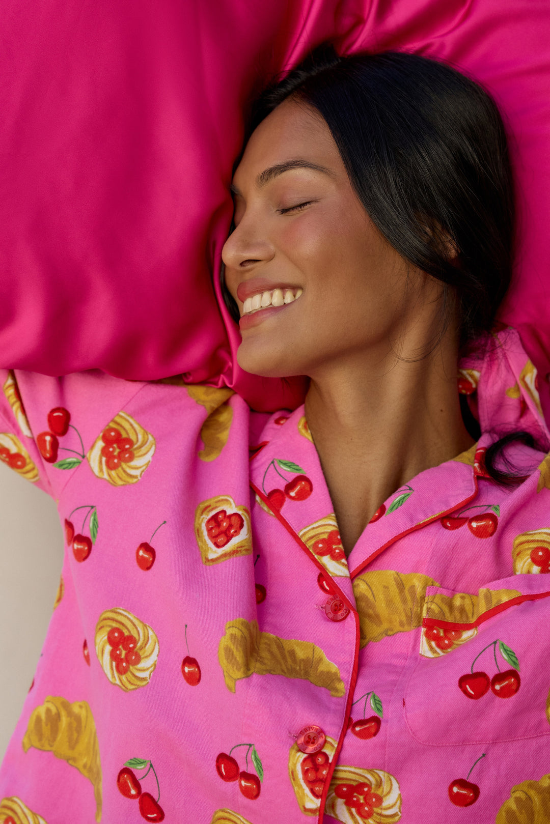 A woman lying down with her eyes closed, wearing bright pink pajamas with a colorful fruit and pastry print, resting on a matching pink pillow.