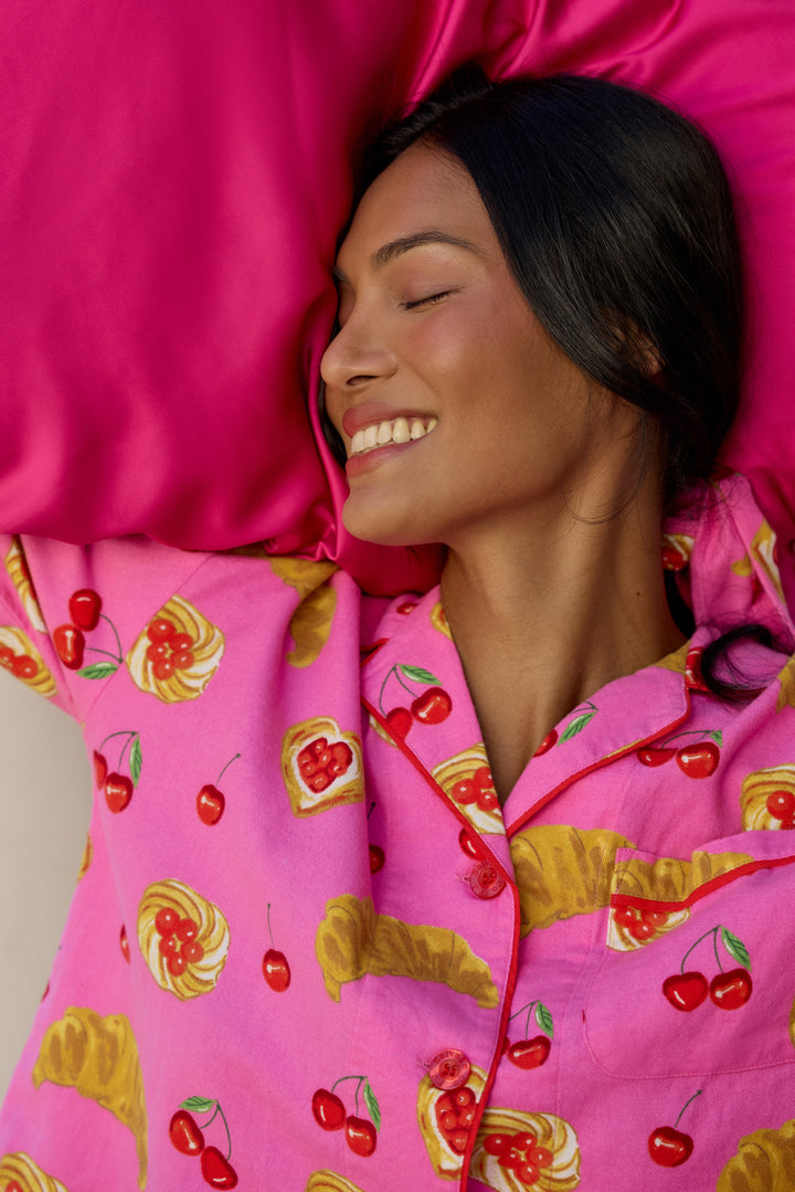 A woman lying down with her eyes closed, wearing bright pink pajamas with a colorful fruit and pastry print, resting on a matching pink pillow.