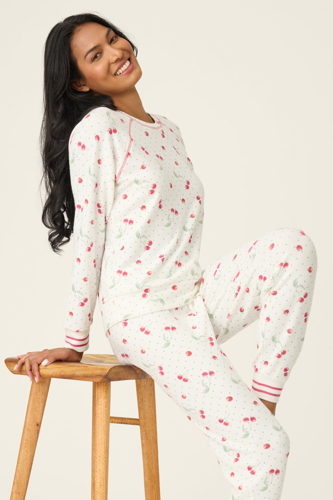 Woman wearing a white pajamas with red cherry pattern, standing on a wooden stool against a plain background.