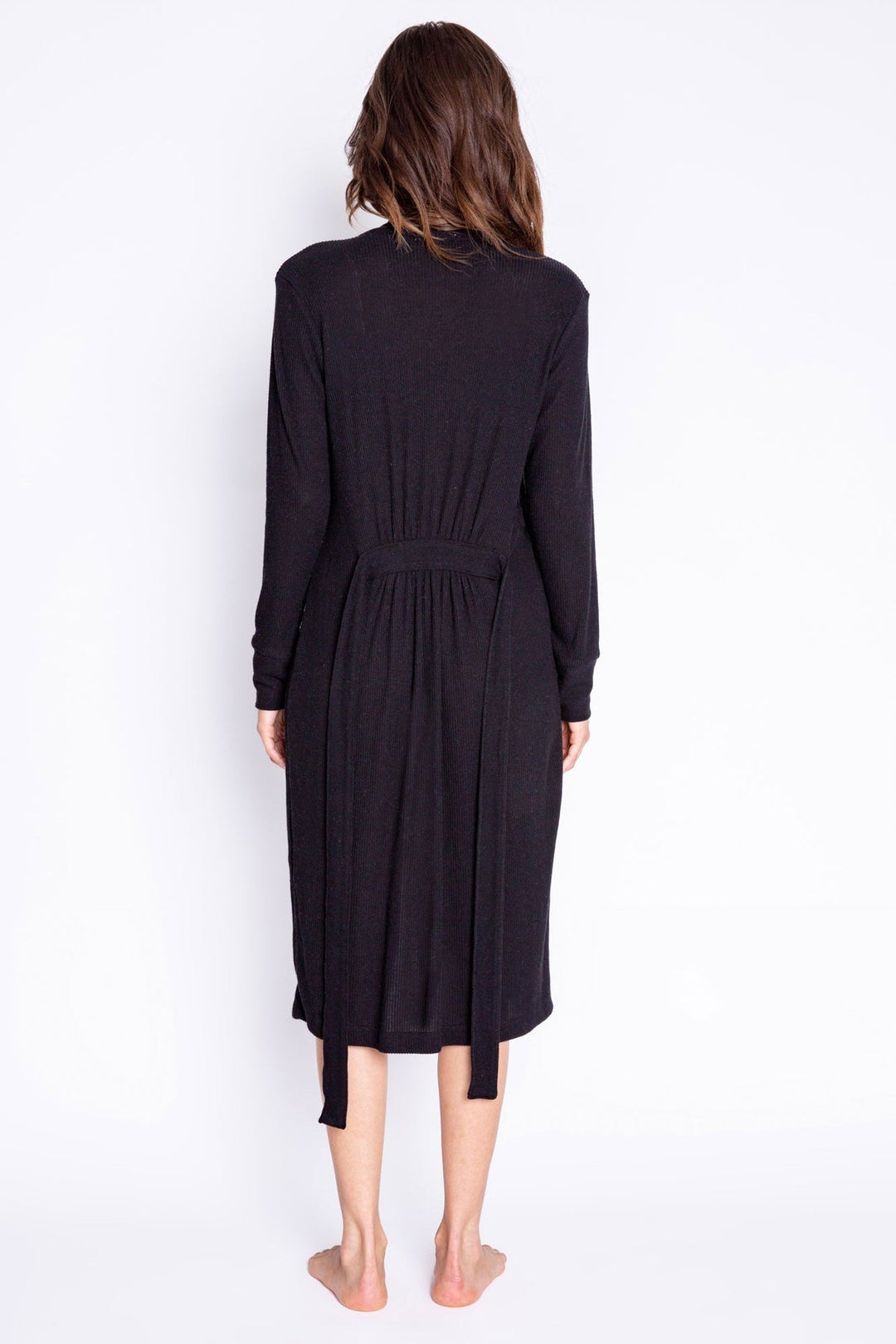 A woman viewed from behind wearing a long-sleeve black midi robe with a tie belt at the waist, standing barefoot against a plain white background.