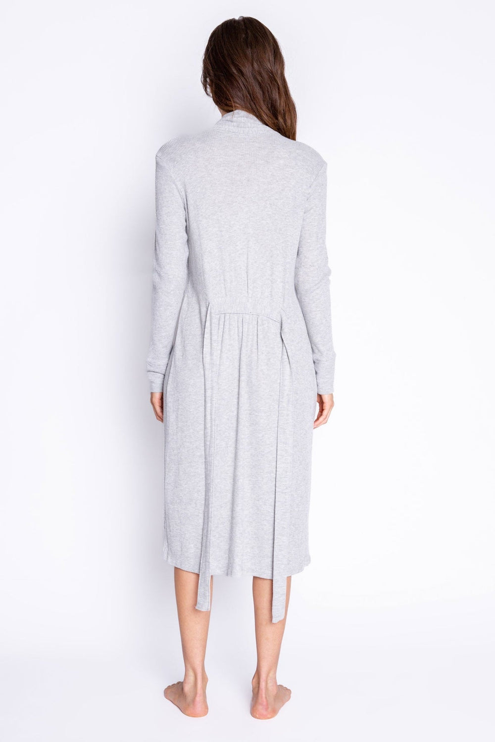 A woman viewed from the back wearing a light gray long-sleeve knit robe with a tie waist, standing barefoot against a plain white studio background.